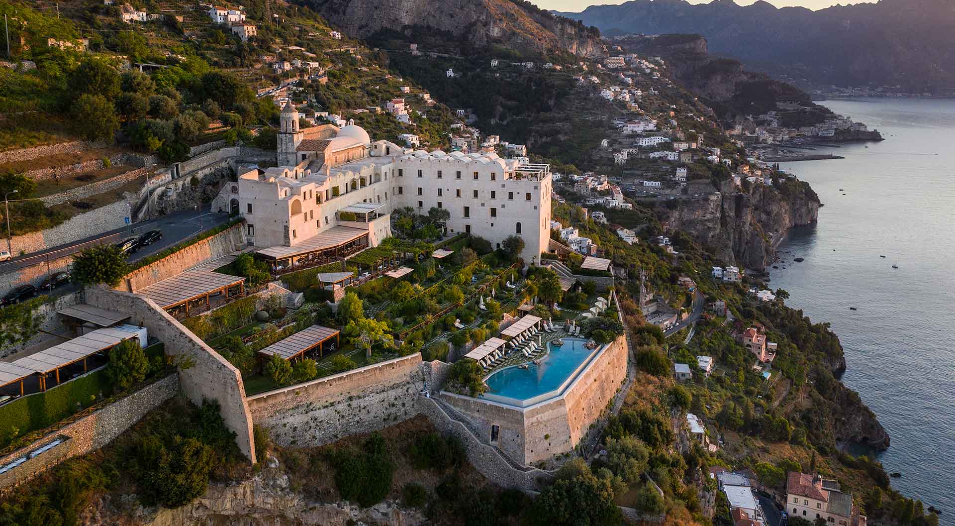 Aerial view of the Monastero Santa Rosa, a luxury hotel perched on a cliff with terraced gardens and an infinity pool overlooking the Amalfi Coast.