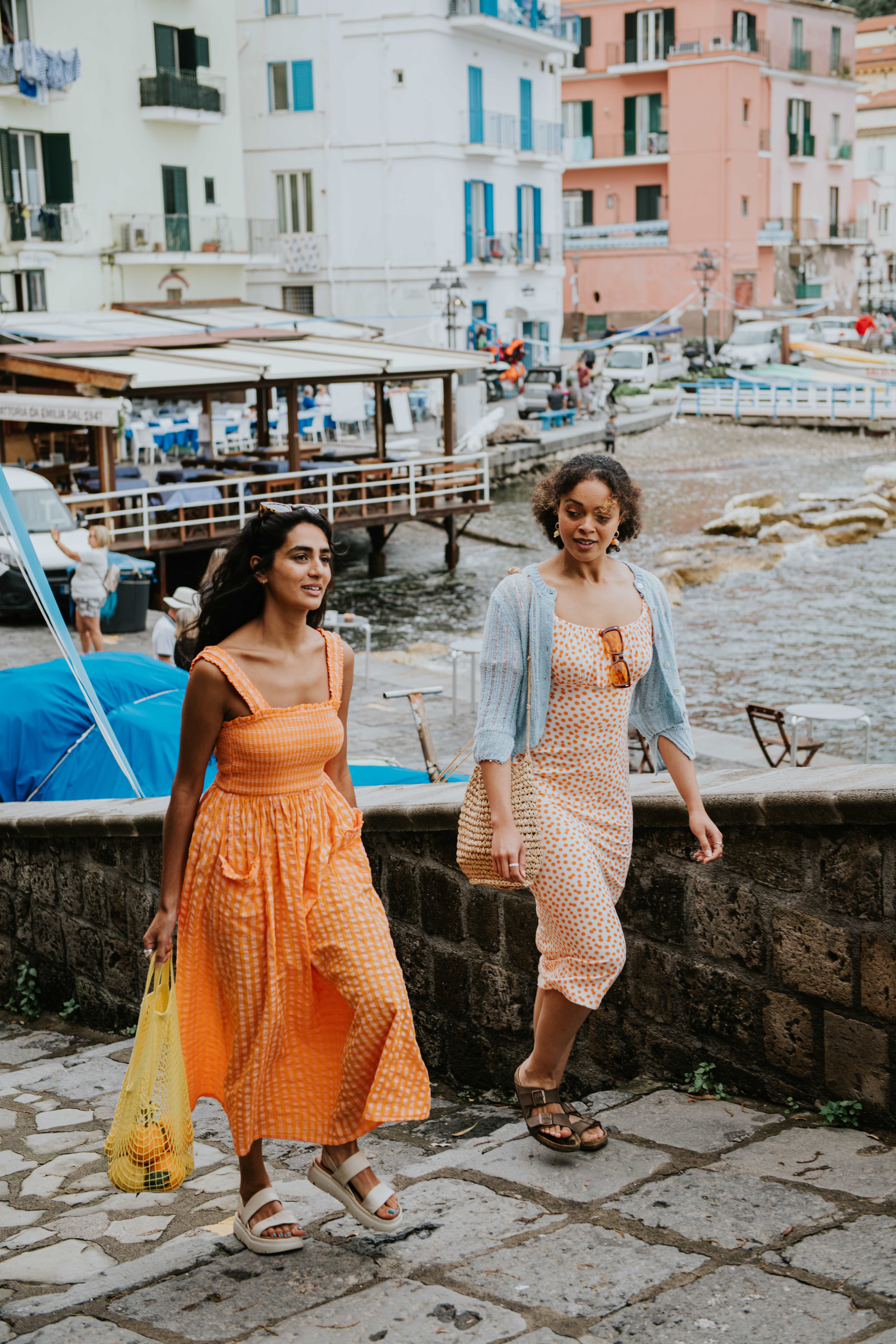 Two women in summer dresses walking along a seaside promenade in a colorful coastal town.