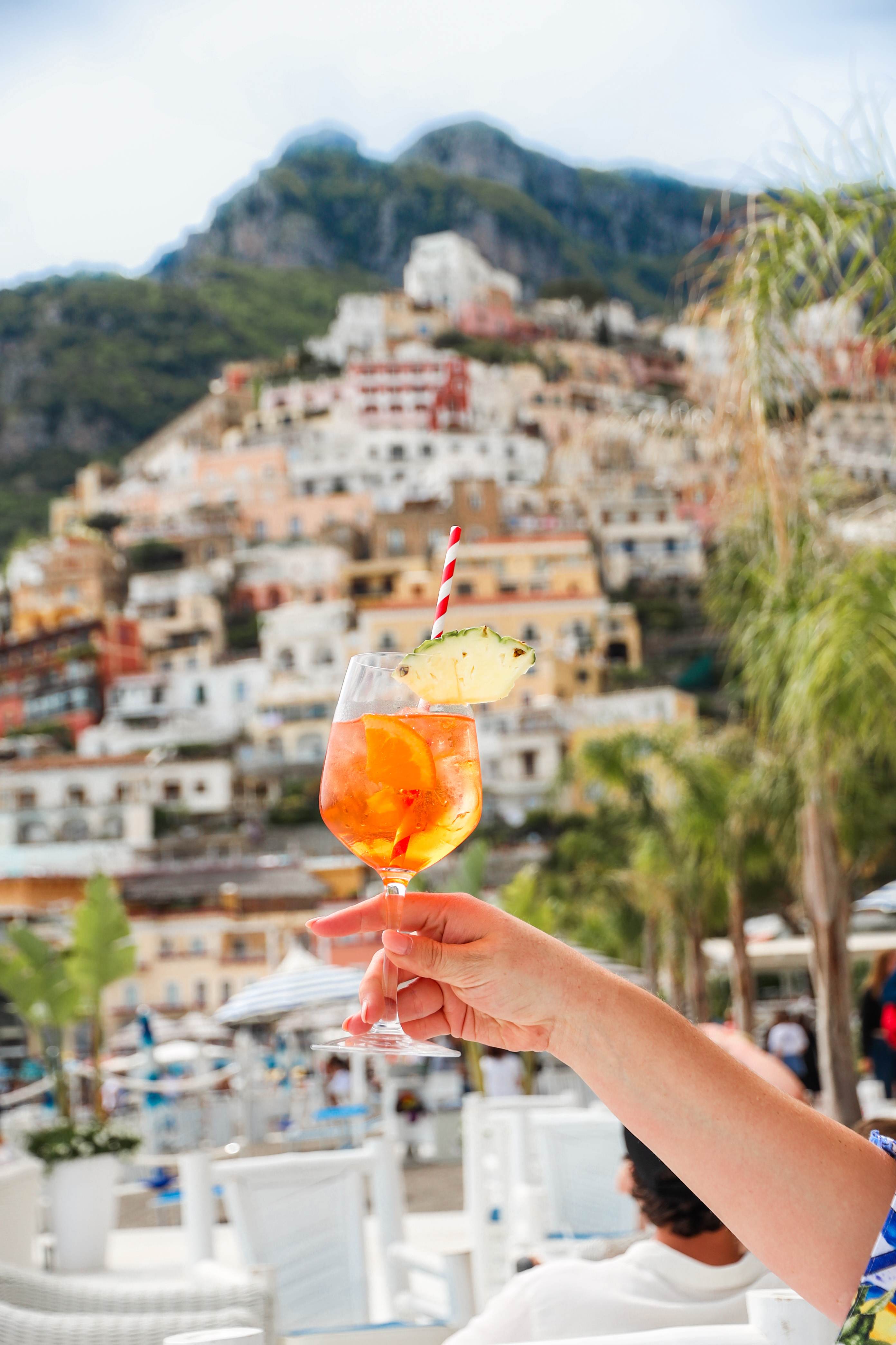 Hand holding a cocktail with an orange slice and a straw, with the colorful hillside buildings of Positano in the background.
