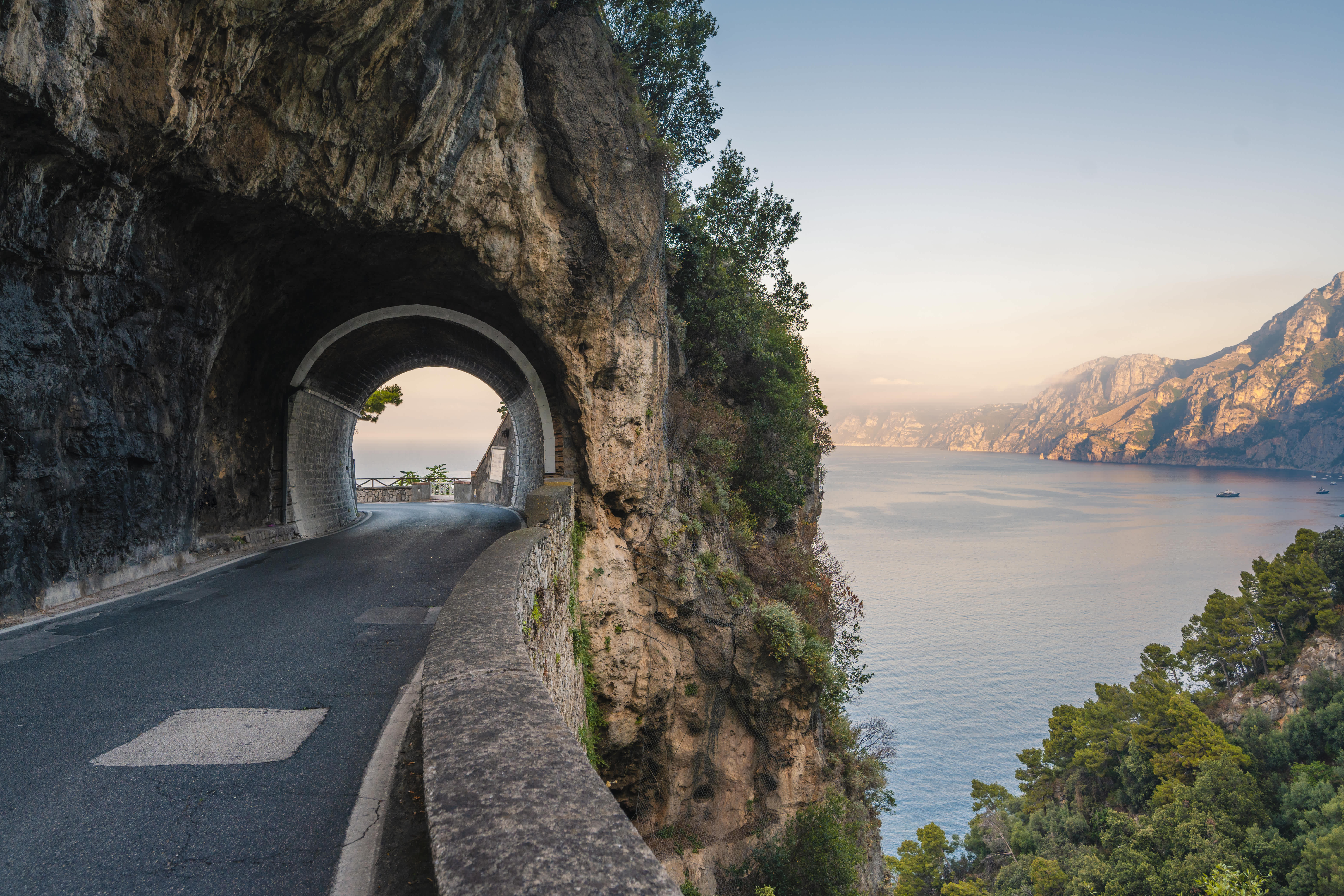 Cliffside road tunnel on the Amalfi Coast with a view of the sea and mountains at sunset.