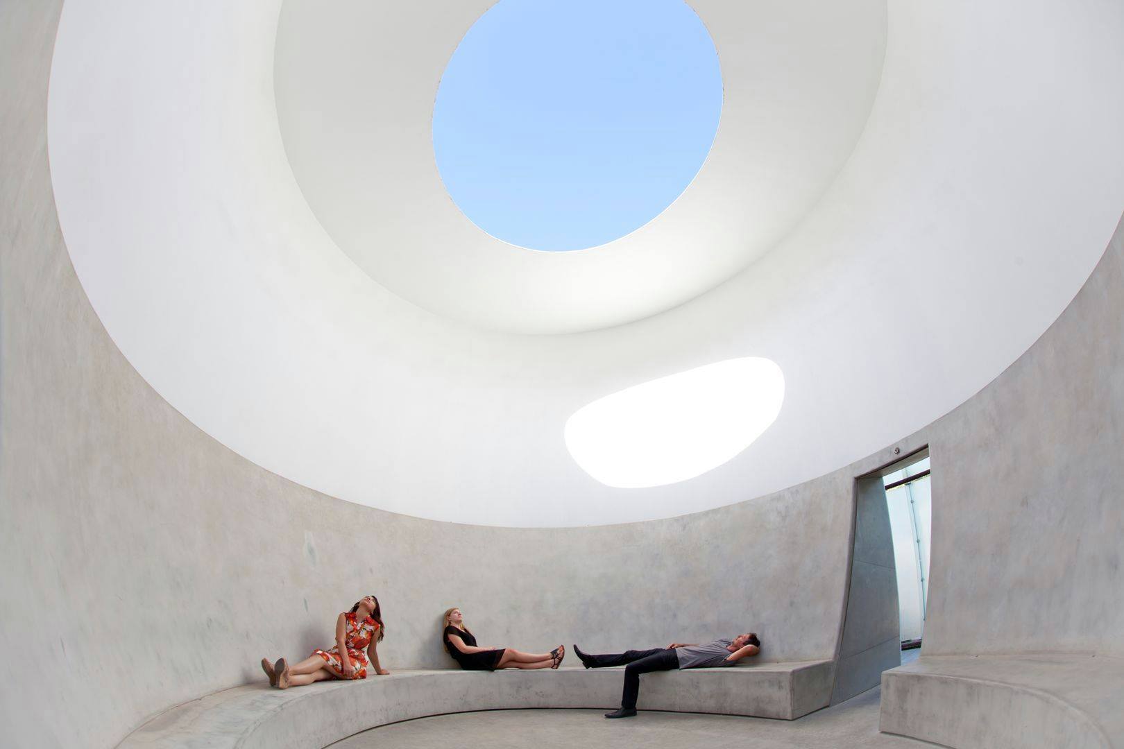 People relaxing inside a modern architectural structure with a large circular opening in the ceiling.