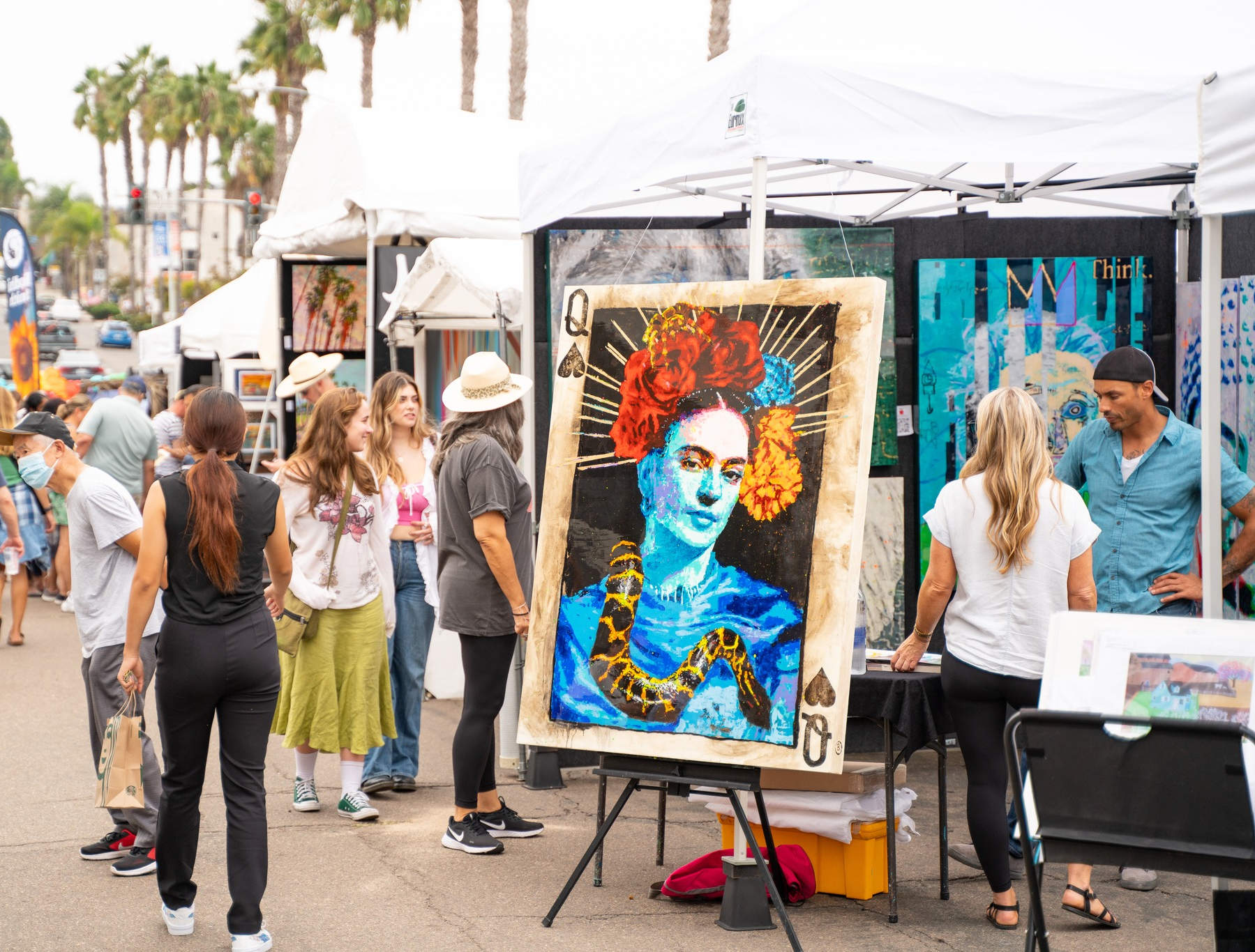 Outdoor art market with several people browsing artwork displayed on white tents and panels.