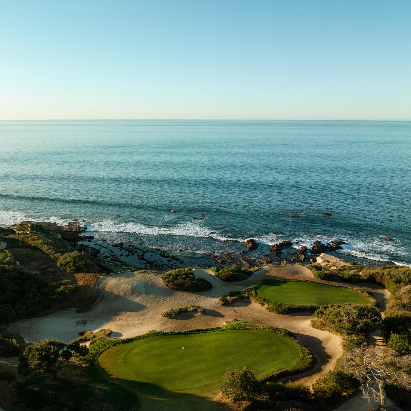 Aerial view of a coastal golf course in Laguna Beach with green fairways, sandy bunkers, and the ocean waves meeting the rocky shoreline.