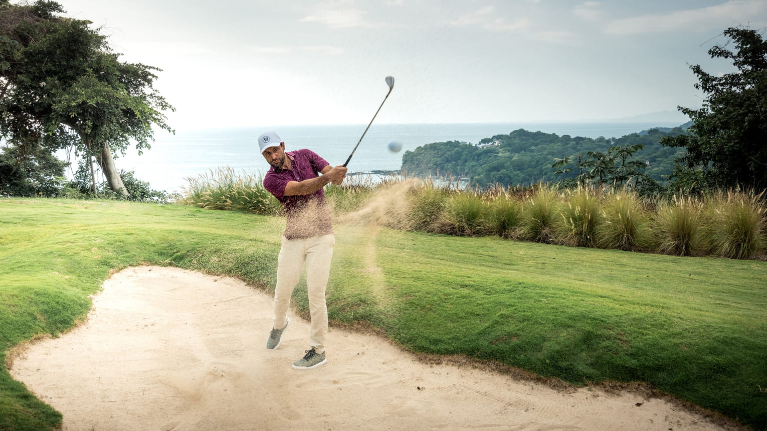 Golfer hitting a shot from a sand bunker with a scenic ocean view in the background.