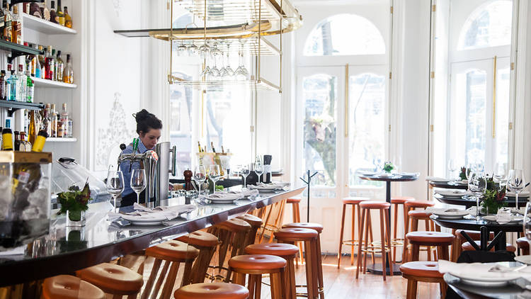 A well-lit restaurant bar with wooden stools, set tables, and a bartender preparing drinks.