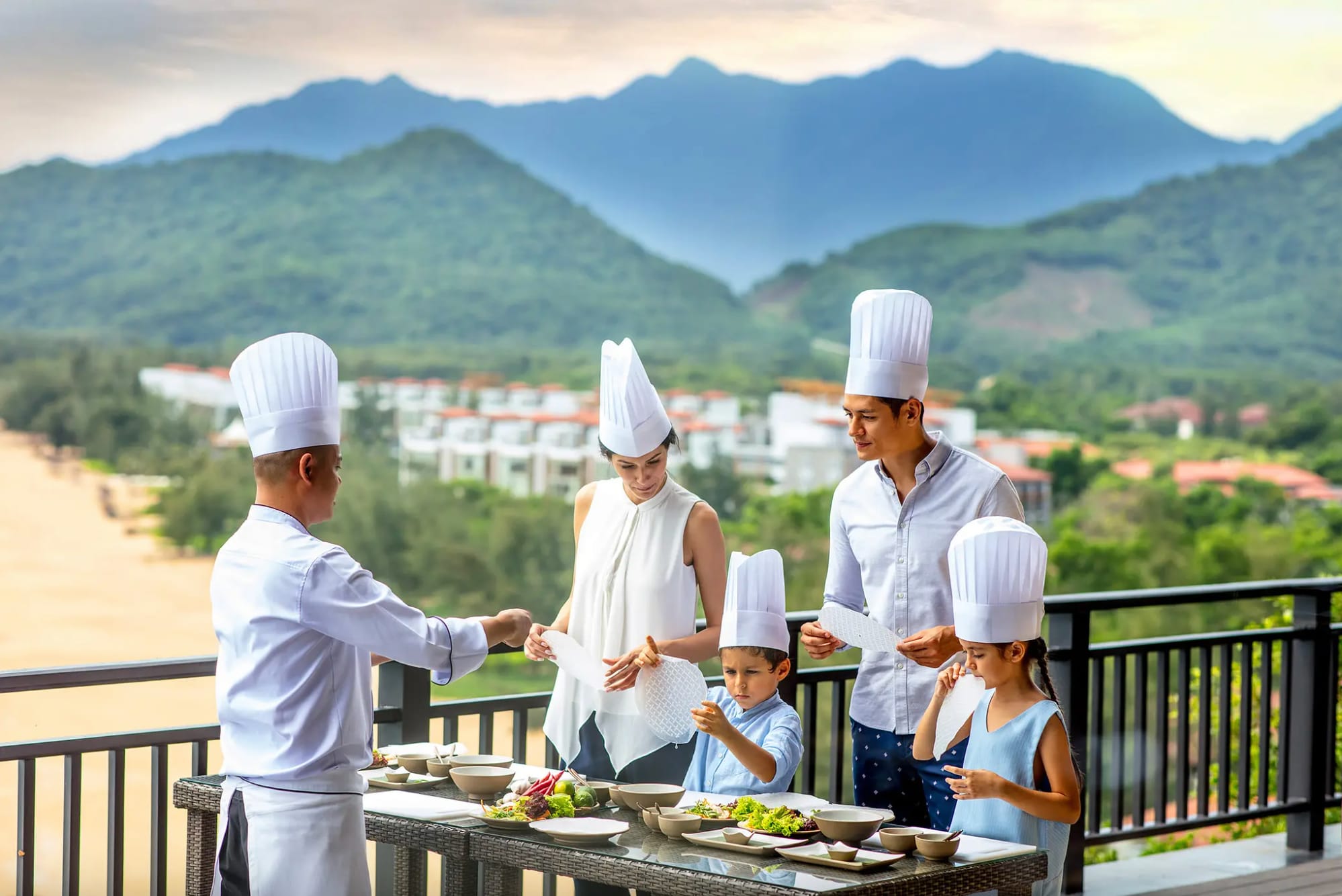 Family learning to cook with a chef on a terrace with mountain views at Banyan Tree Lang Co, Vietnam.