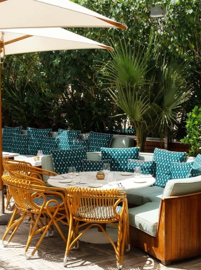 Outdoor dining area with round tables, wicker chairs, and teal cushions surrounded by greenery.