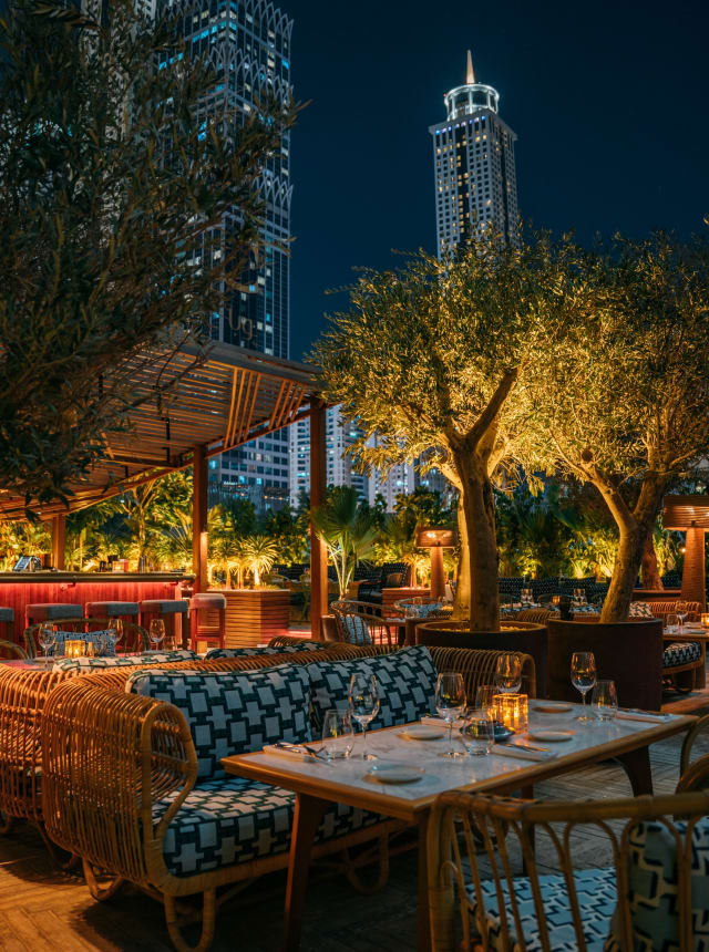 Outdoor restaurant at night with cozy seating, ambient lighting, and trees, set against the backdrop of tall city buildings.