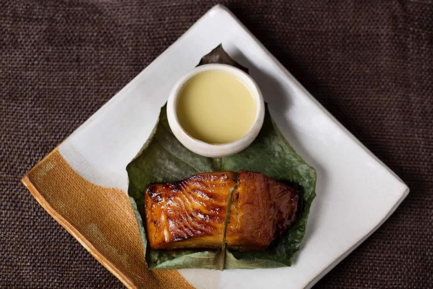 Grilled miso-marinated black cod served on a leaf with a side of sauce at Zuma restaurant.
