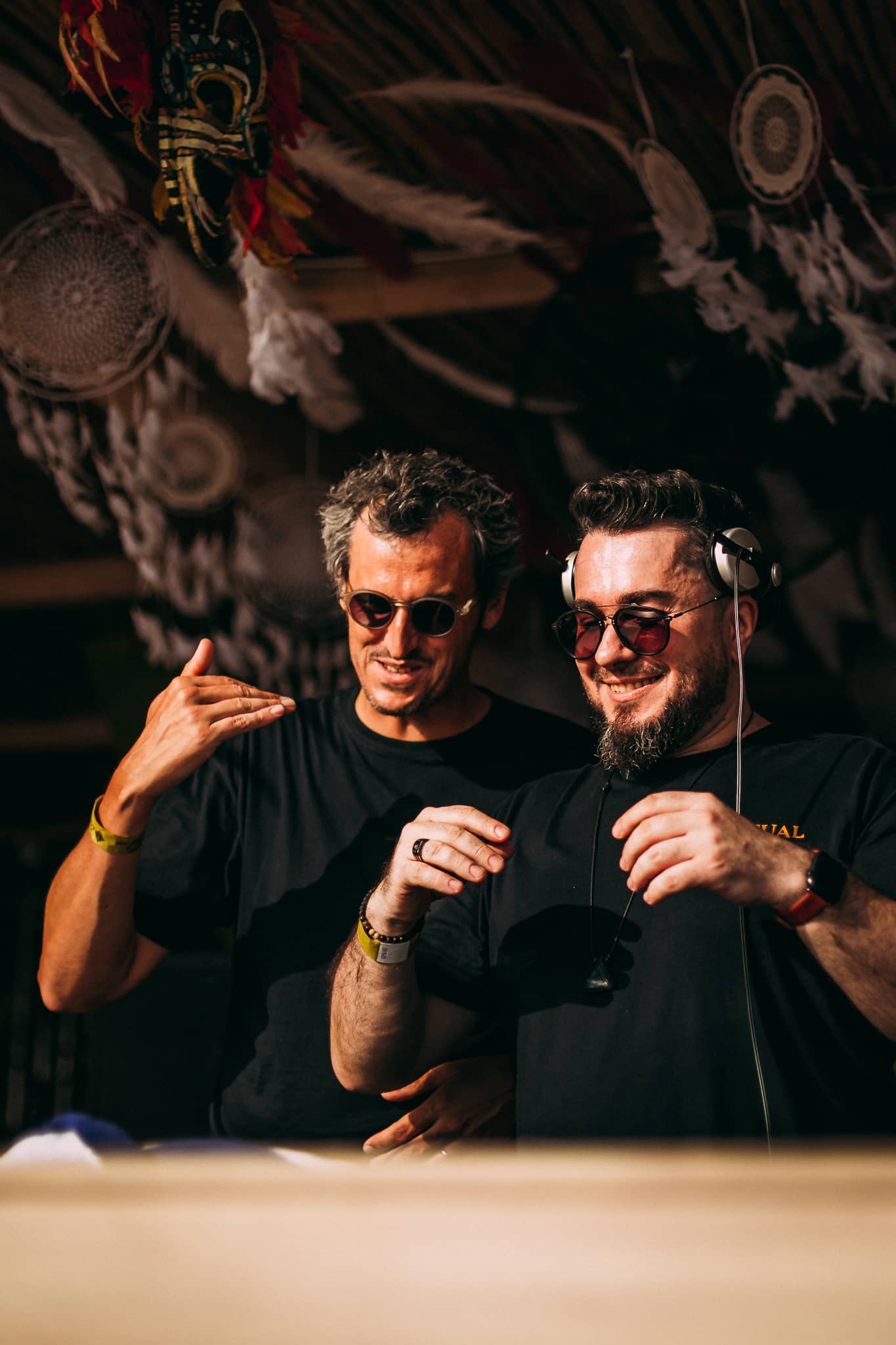 Two DJs wearing sunglasses performing together at an outdoor event, with dreamcatchers hanging in the background.