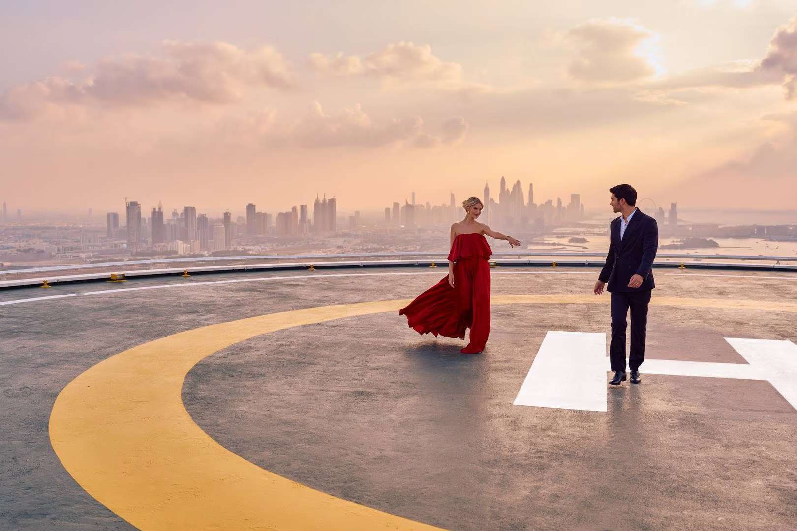 Couple elegantly dressed on a helipad with a panoramic view of the Dubai skyline at sunset.