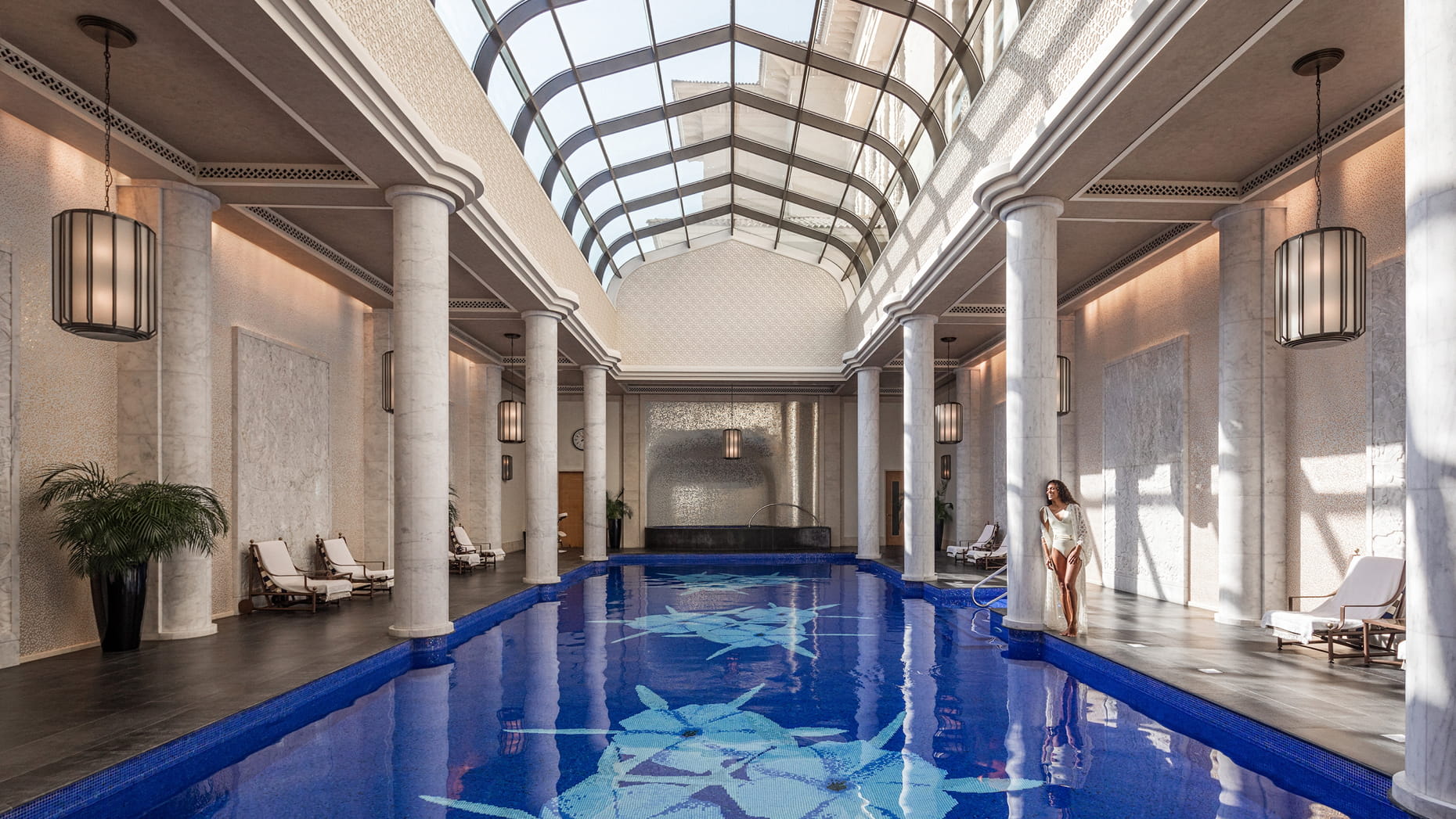 Elegant indoor pool area with marble columns, large skylights, and lounge chairs, with a woman standing by the pool.