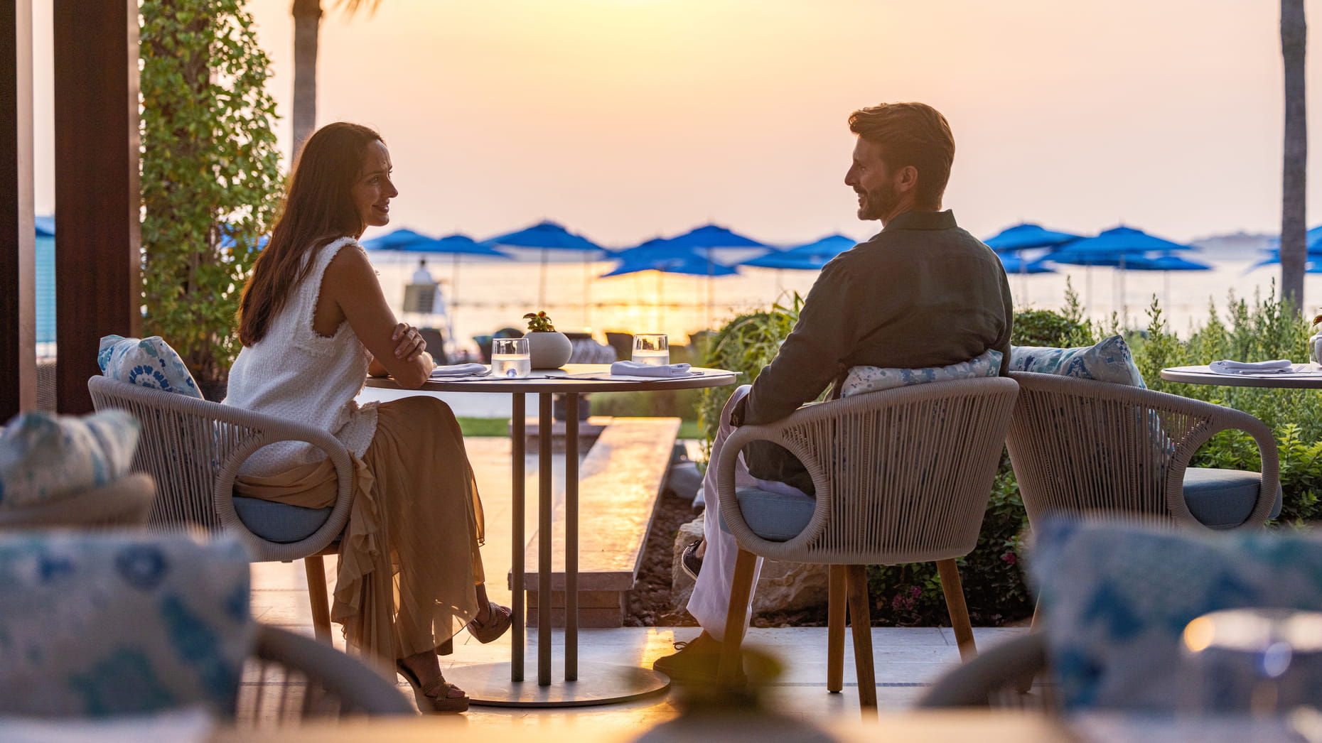 Couple enjoying a sunset dinner outdoors with a view of the beach and blue umbrellas in the background.