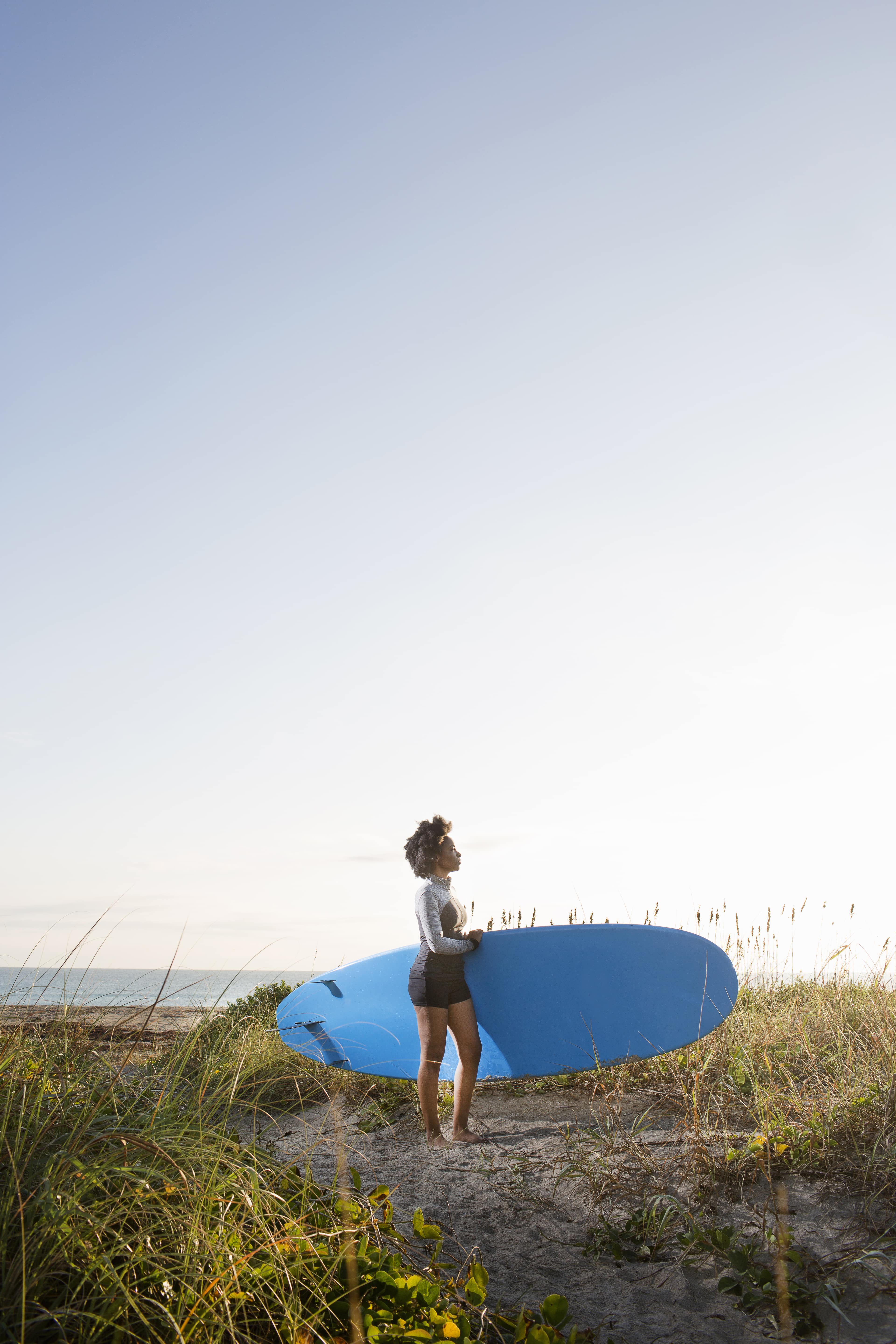 Woman standing on a sandy path holding a blue surfboard, looking out over the ocean at sunrise on Kiawah Island.