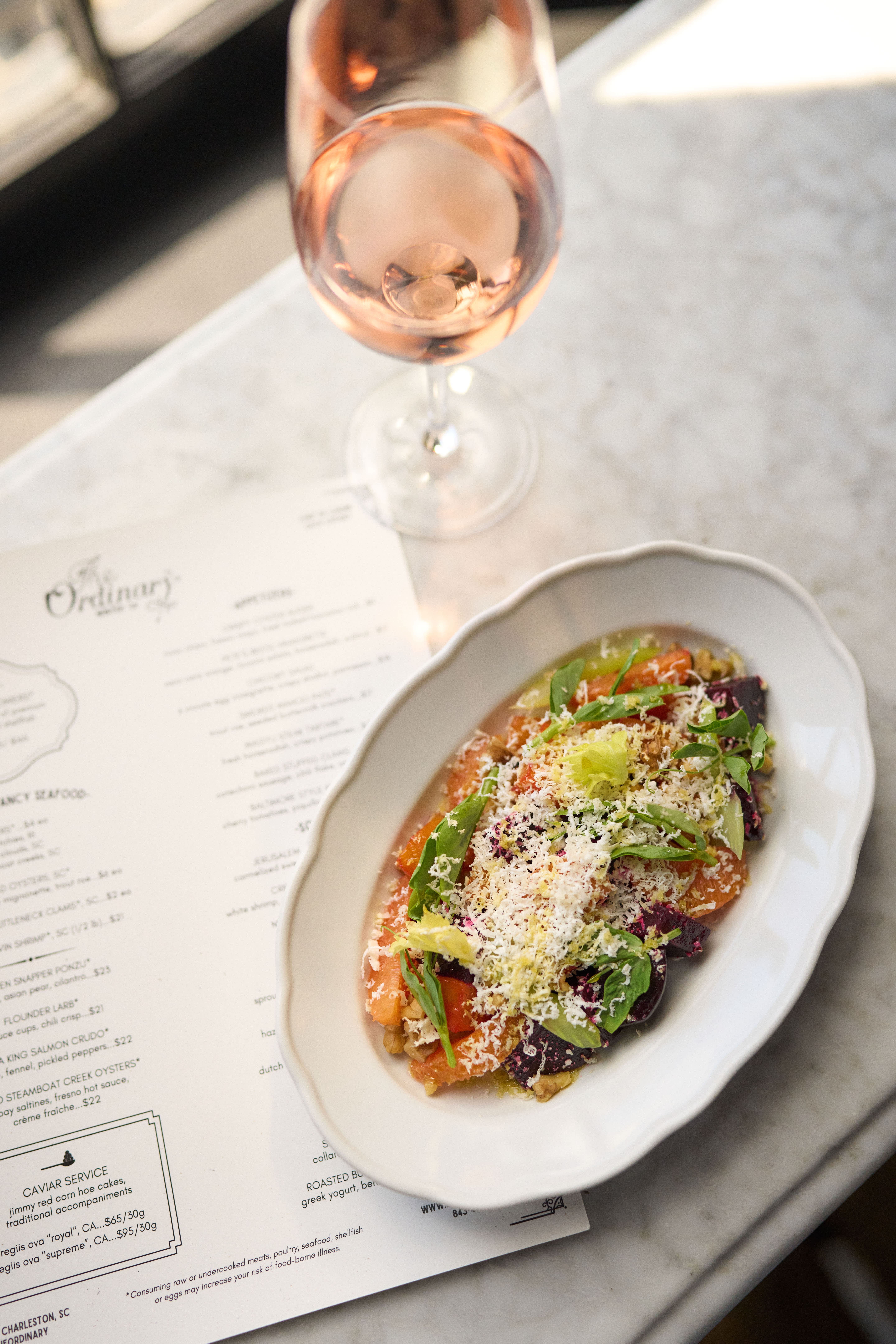 Glass of rosé wine on a marble table next to a vibrant salad with fresh greens, tomatoes, and cheese, with The Ordinary's menu in the background.