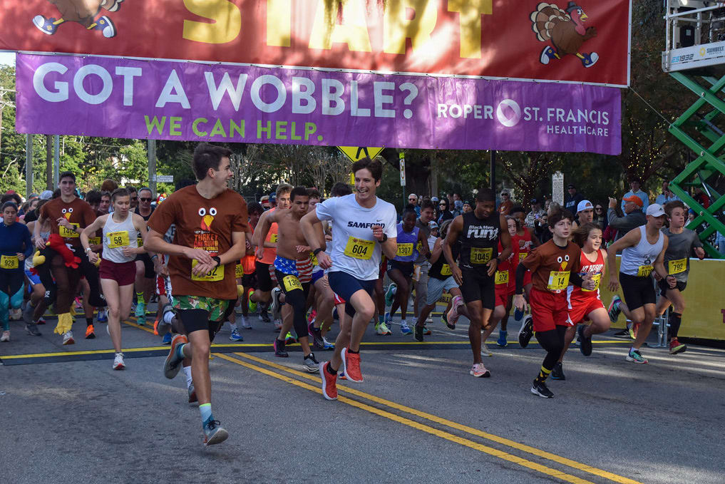 Runners dressed in turkey-themed outfits and athletic gear take off at the start of a festive race, beneath a banner promoting the event.