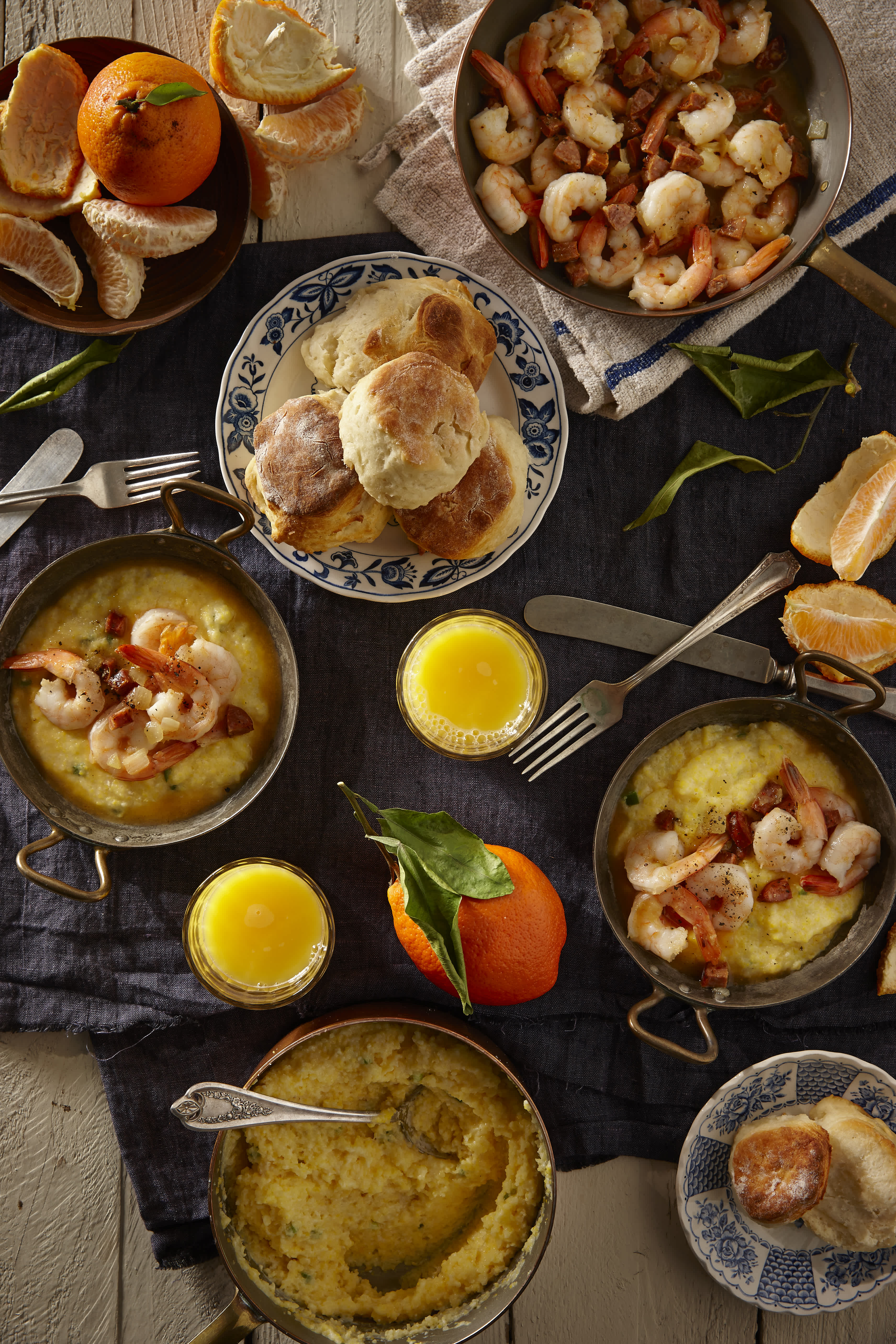 Top-down view of a Southern breakfast spread featuring shrimp and grits, freshly baked biscuits, oranges, and glasses of orange juice.