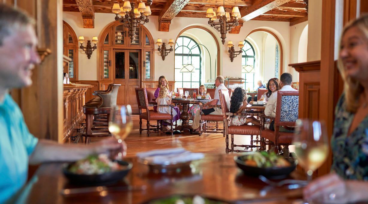 Guests dining in an elegant, wood-paneled restaurant with large windows and chandeliers, enjoying meals and conversation at their tables.