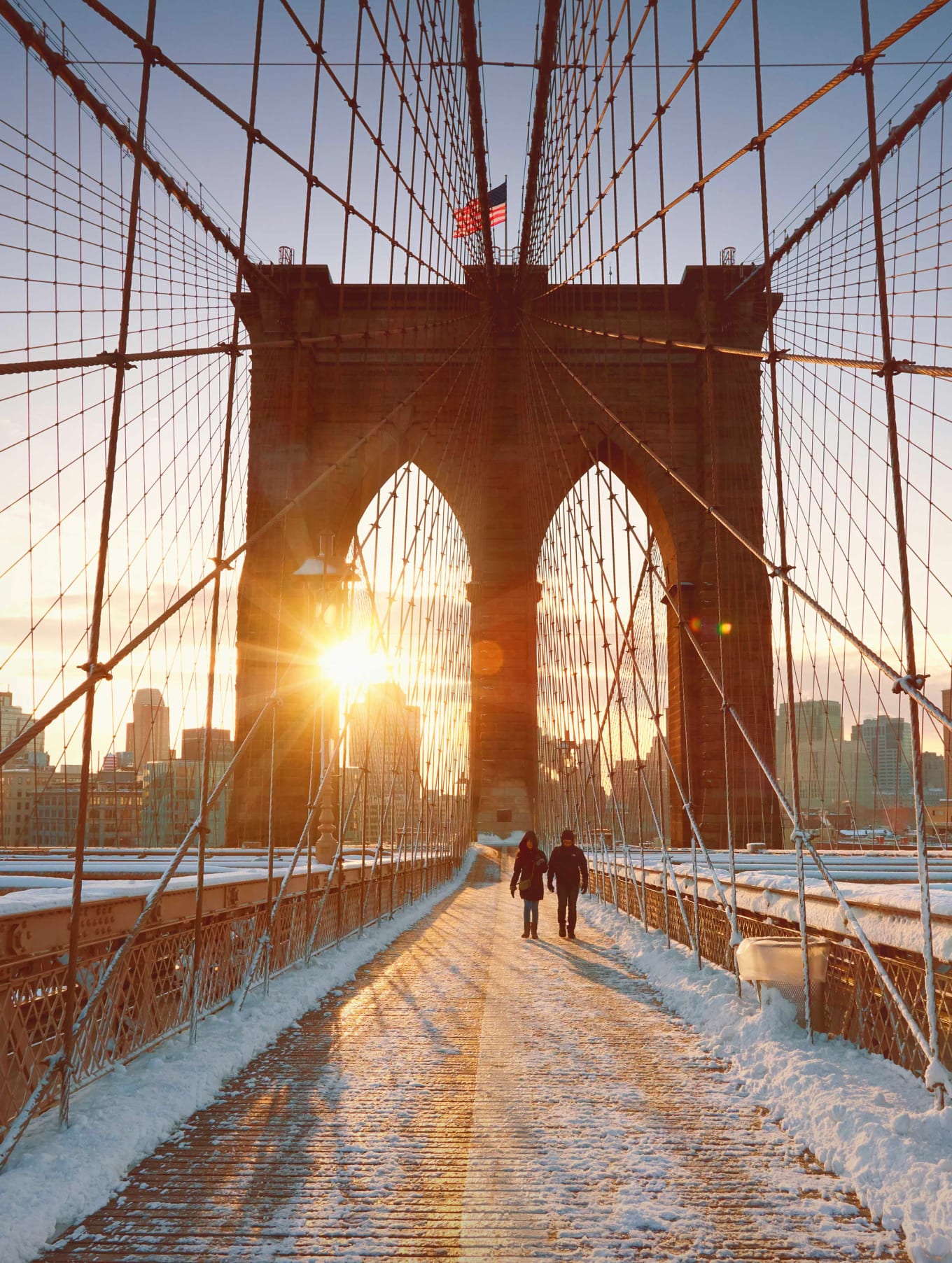 Winter sunrise casting over the snow-covered Brooklyn Bridge with pedestrians strolling along the pathway.